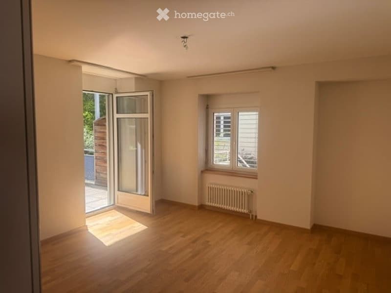 Empty room with wooden flooring, white walls, and a radiator under a window. A sliding glass door leads to an outdoor area.