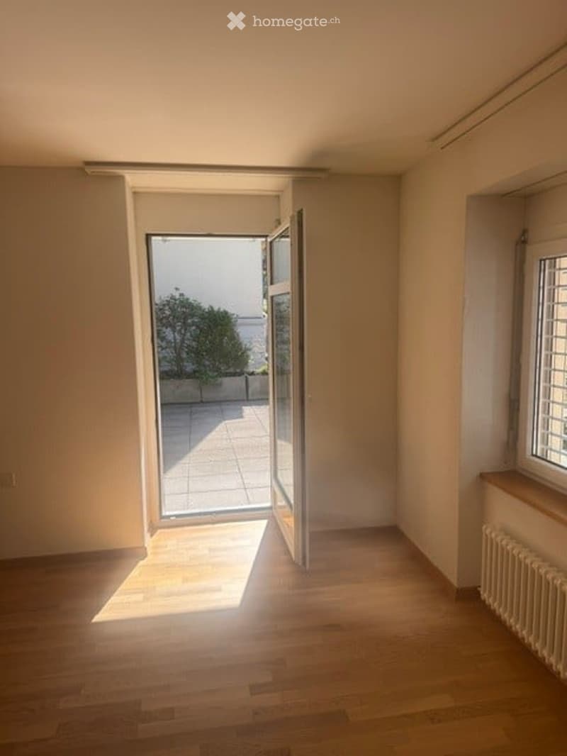 Empty room with wooden flooring, large window, and open sliding door leading to a patio area.