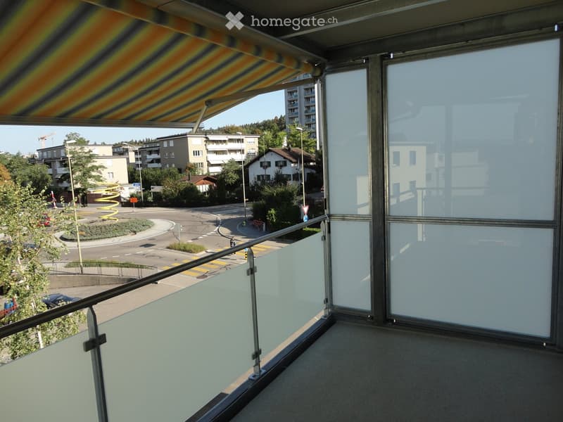 Balcony with glass railings, overlooking a street with buildings, trees, and traffic.