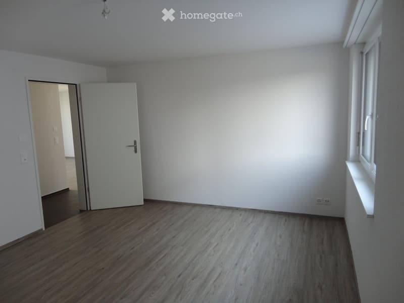 Empty room with light-colored walls, wooden flooring, and a window with a white sill. A closed door leads to another space.
