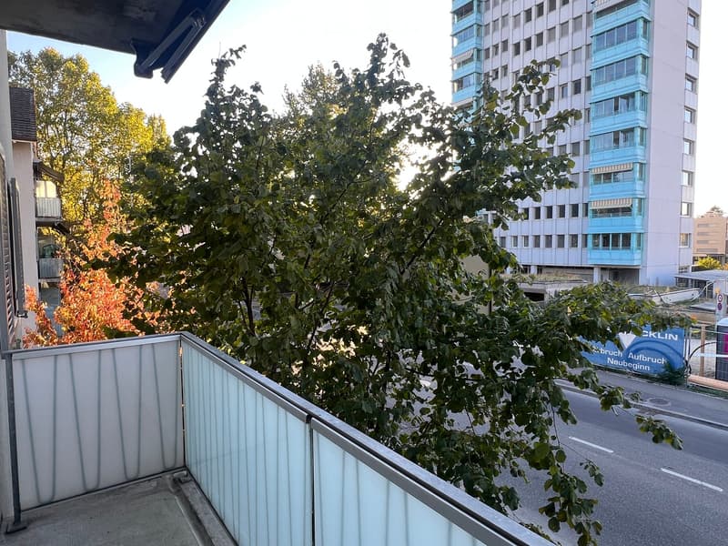 Balcony with a metal railing, overlooking a street with trees and a multi-story building in the background.
