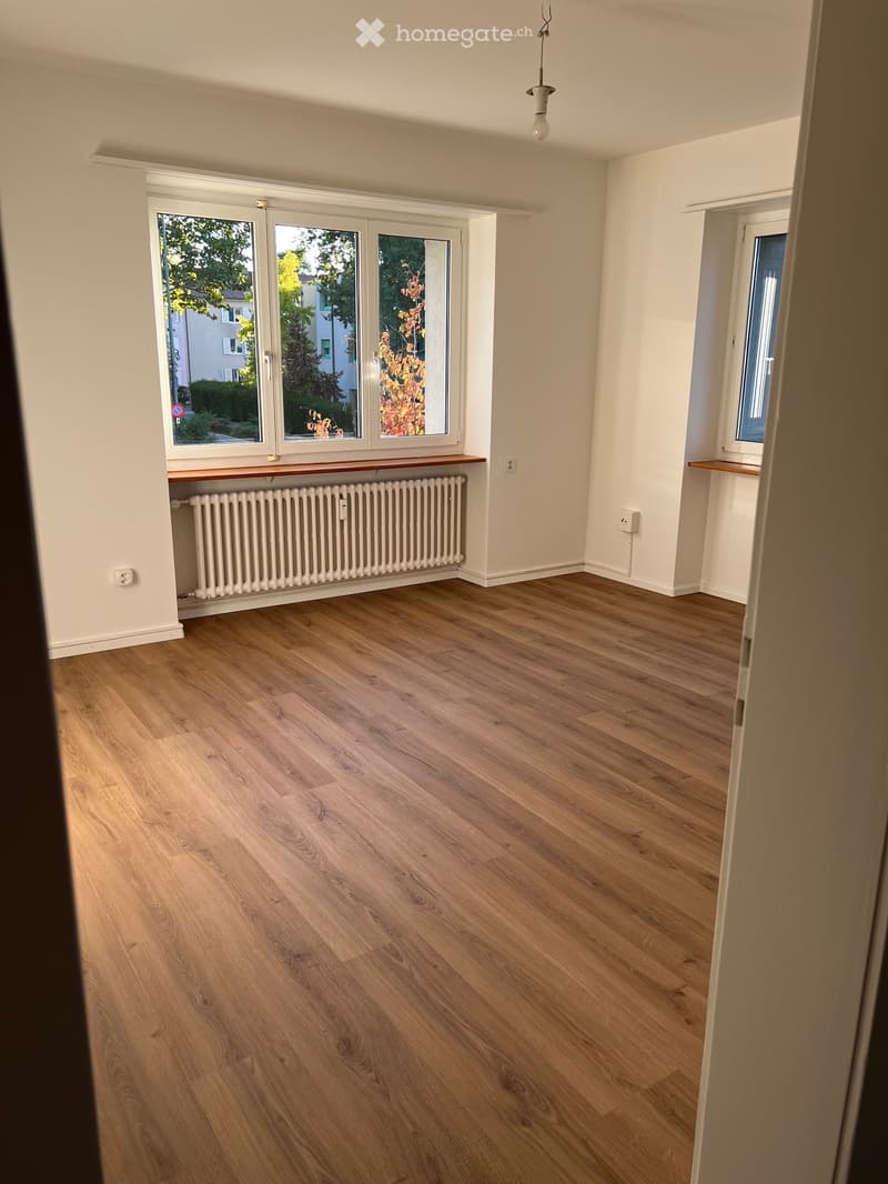 Empty room with wooden flooring, white walls, large windows, radiator, and a small shelf.