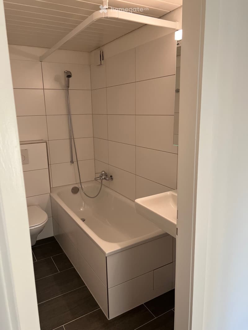 Modern bathroom with a bathtub, toilet, sink, and showerhead. The walls are tiled in light beige, and the floor is dark grey.