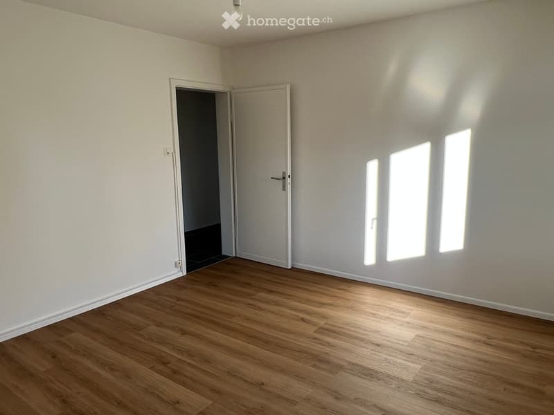 Empty room with wooden flooring, white walls, and large windows allowing natural light. Features a closed door and an open doorway leading to another space.