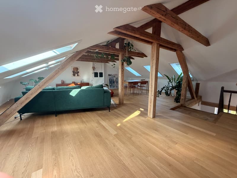 Open-plan attic space with wooden beams, skylights, hardwood flooring, green sofa, dining table, chairs, and artwork.