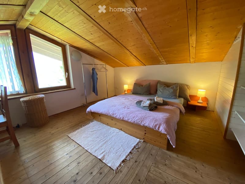 Cozy attic bedroom with wooden slanted ceilings, a large bed with pillows and blankets, nightstands with lamps, a wicker basket, and a clothing rack.