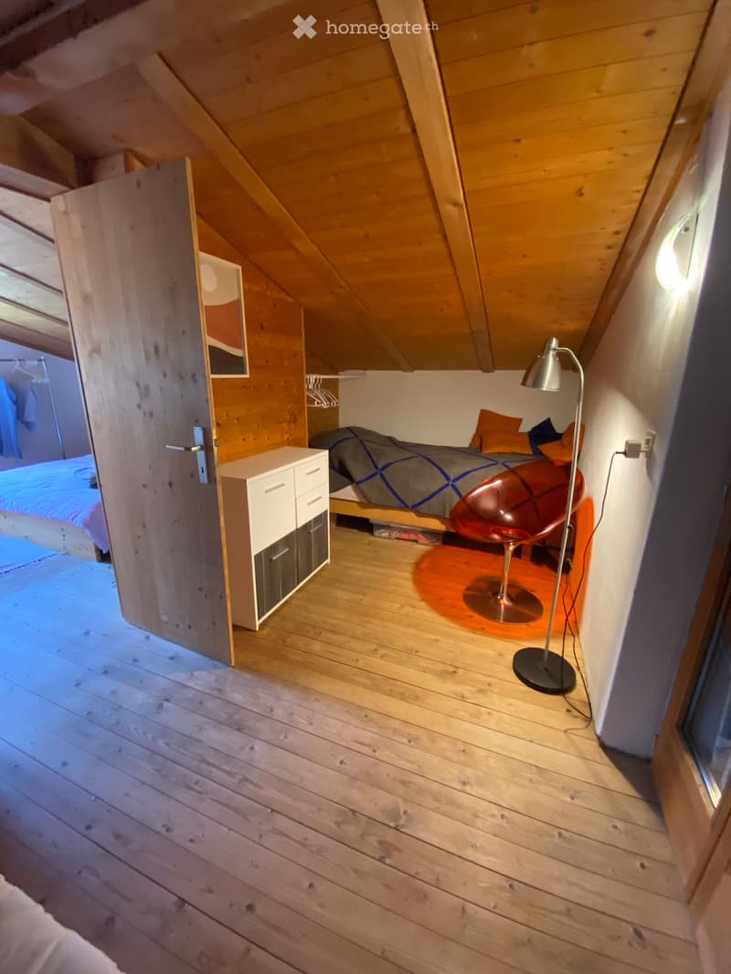 Cozy attic bedroom with wooden walls and floor, featuring a bed with pillows, a red chair, a white cabinet, and a floor lamp.