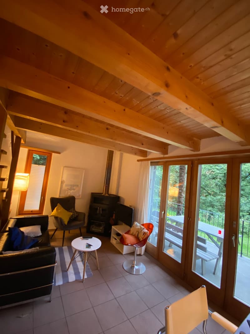 Living room with exposed wooden beams, fireplace, sofa, chairs, small table, and large windows opening to a balcony.