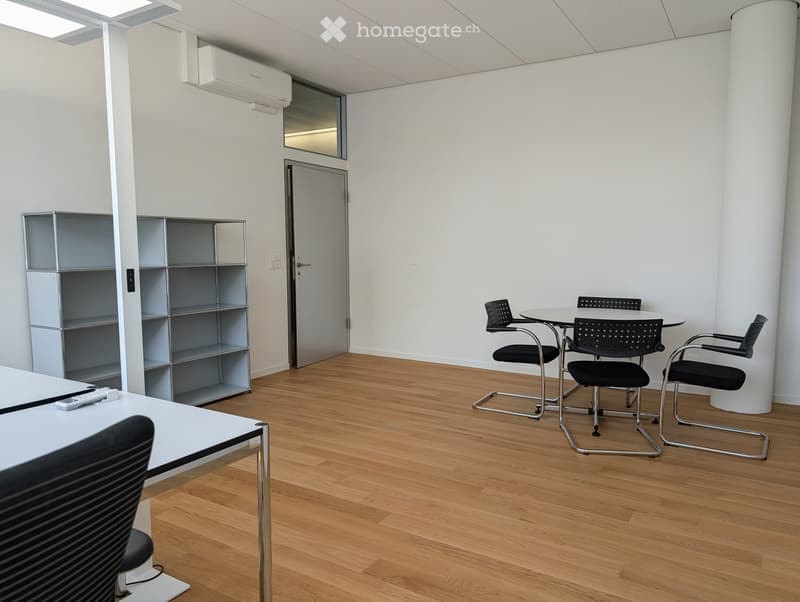 Modern office space with light wooden flooring, white walls, and a large window. Includes a round meeting table with four chairs, a white desk with a chair, and a metal shelving unit.