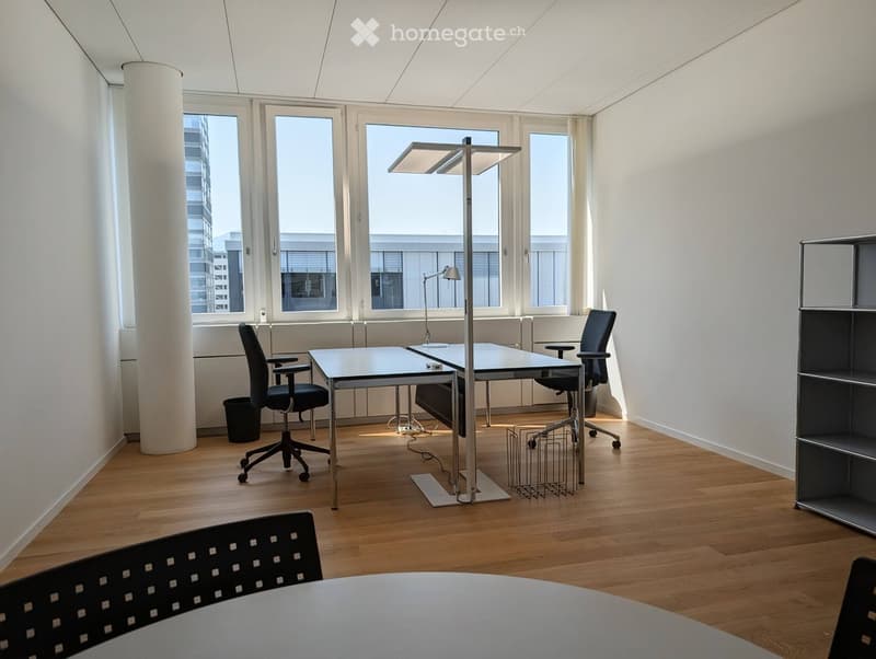 Modern office space with wooden flooring, large windows, a white table, two office chairs, a floor lamp, and a black shelving unit.