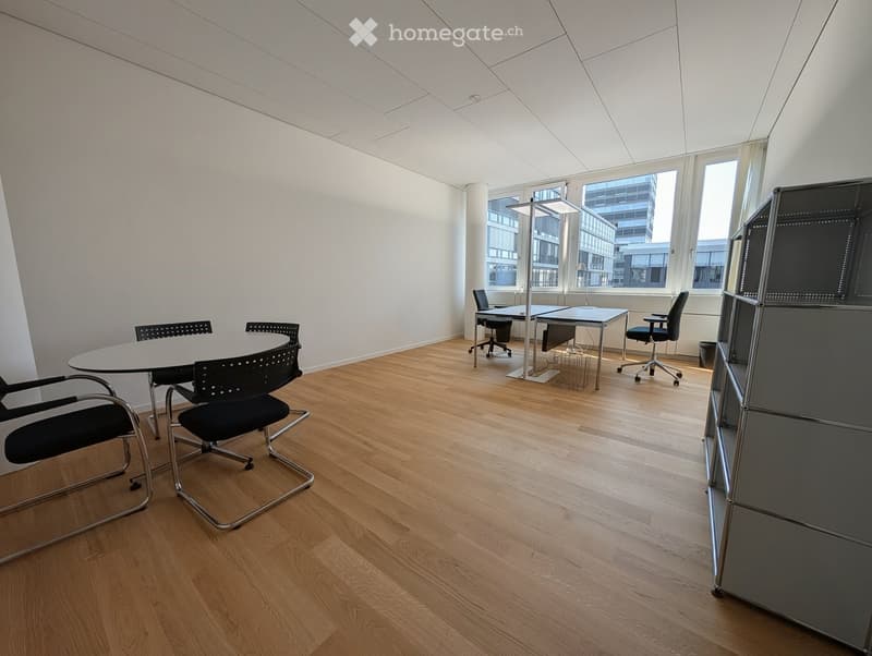 Modern office space with light wooden flooring, large windows, a round meeting table with chairs, a desk with office chair, and a floor lamp.