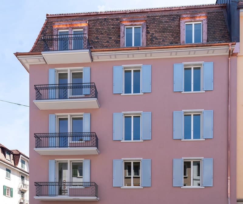 Pink apartment building with multiple balconies and shutters, featuring a tiled roof and classic architectural details.