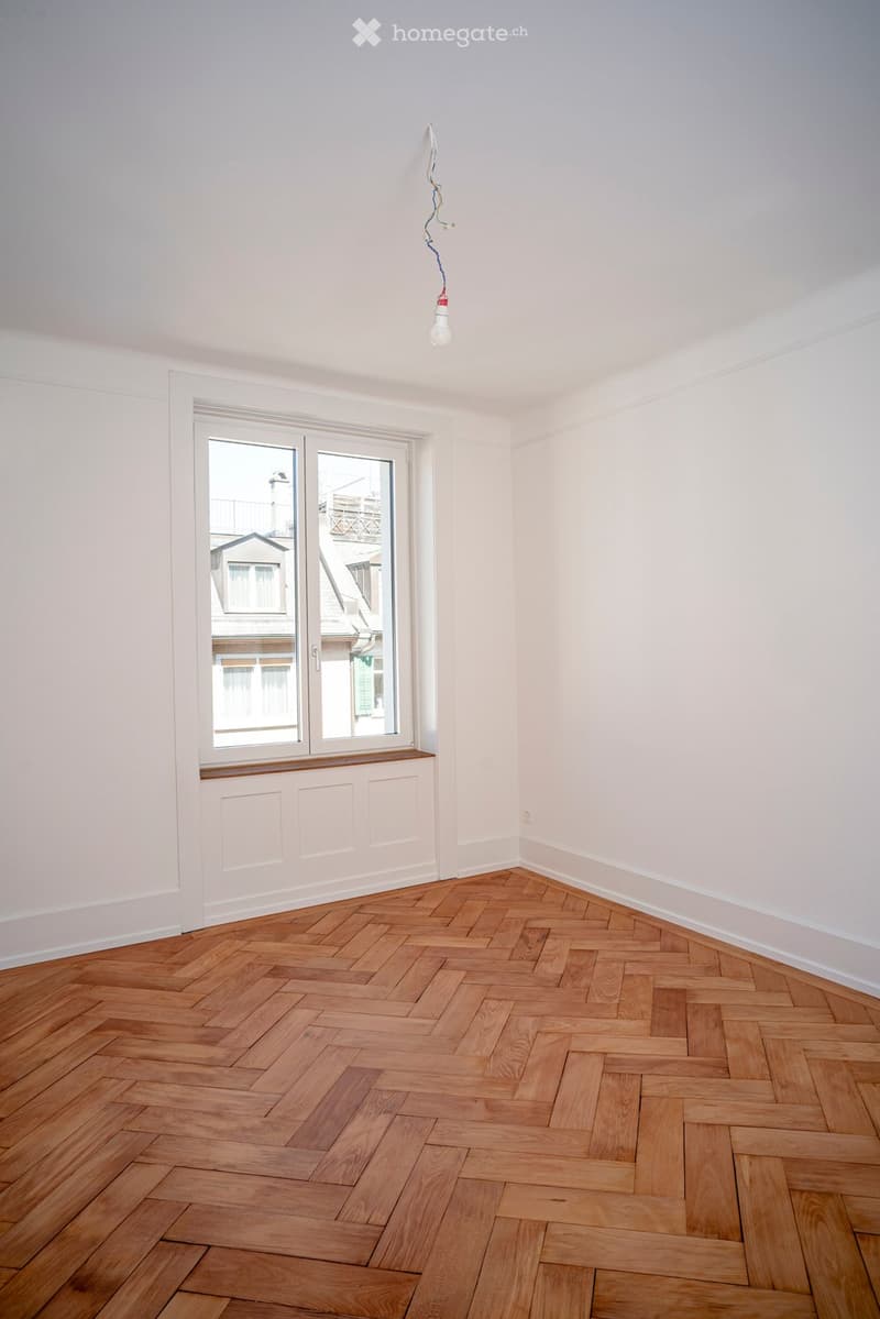 Empty room with light-colored walls, wooden herringbone floor, and a single window with a view of neighboring buildings.