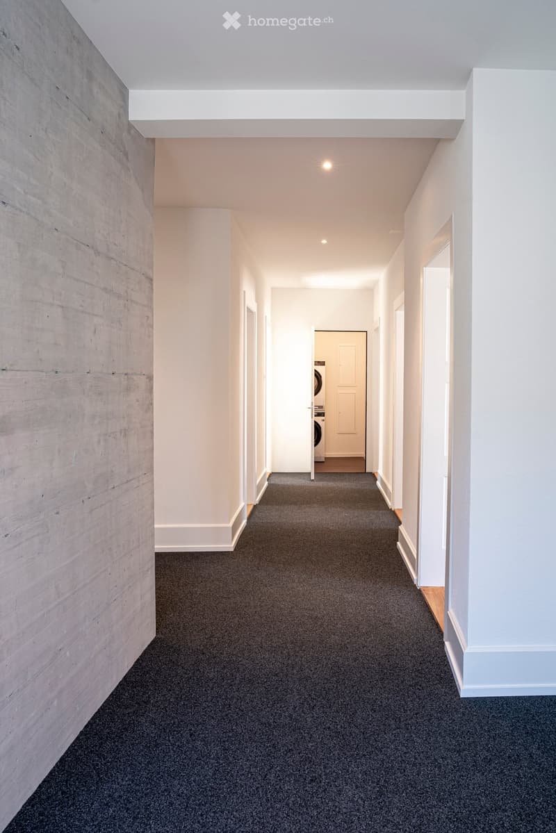 A modern, carpeted hallway with white walls, recessed lighting, and a wooden accent wall on the left side. The hallway leads to a room with a washing machine visible at the end.
