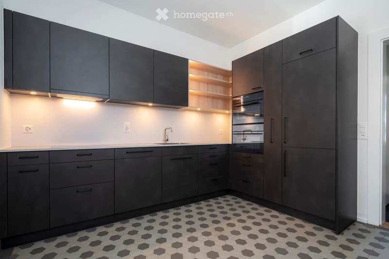 Modern kitchen with dark grey cabinets, white walls, and a hexagonal tile floor. Features include a sink with a modern faucet, built-in lighting, and a built-in oven.