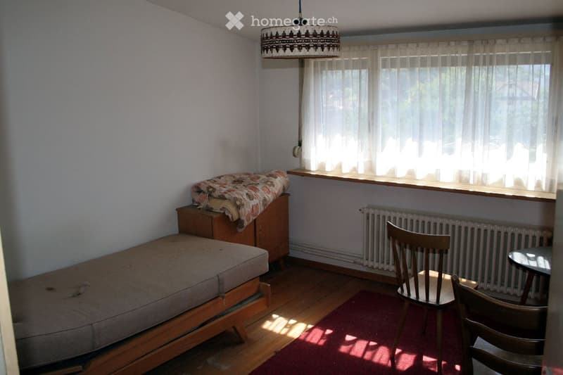 A small, cozy room with wooden flooring, a sofa, a chair, a small table, a radiator, and a window with curtains. A patterned blanket is placed on a wooden cabinet.