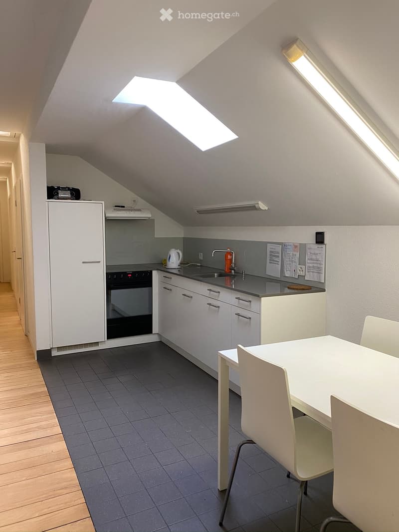 Modern kitchen with white cabinets, stainless steel appliances, and a dining area featuring a white table and chairs. The space has a slanted ceiling with skylights, wooden flooring, and grey tiled flooring in the kitchen area.