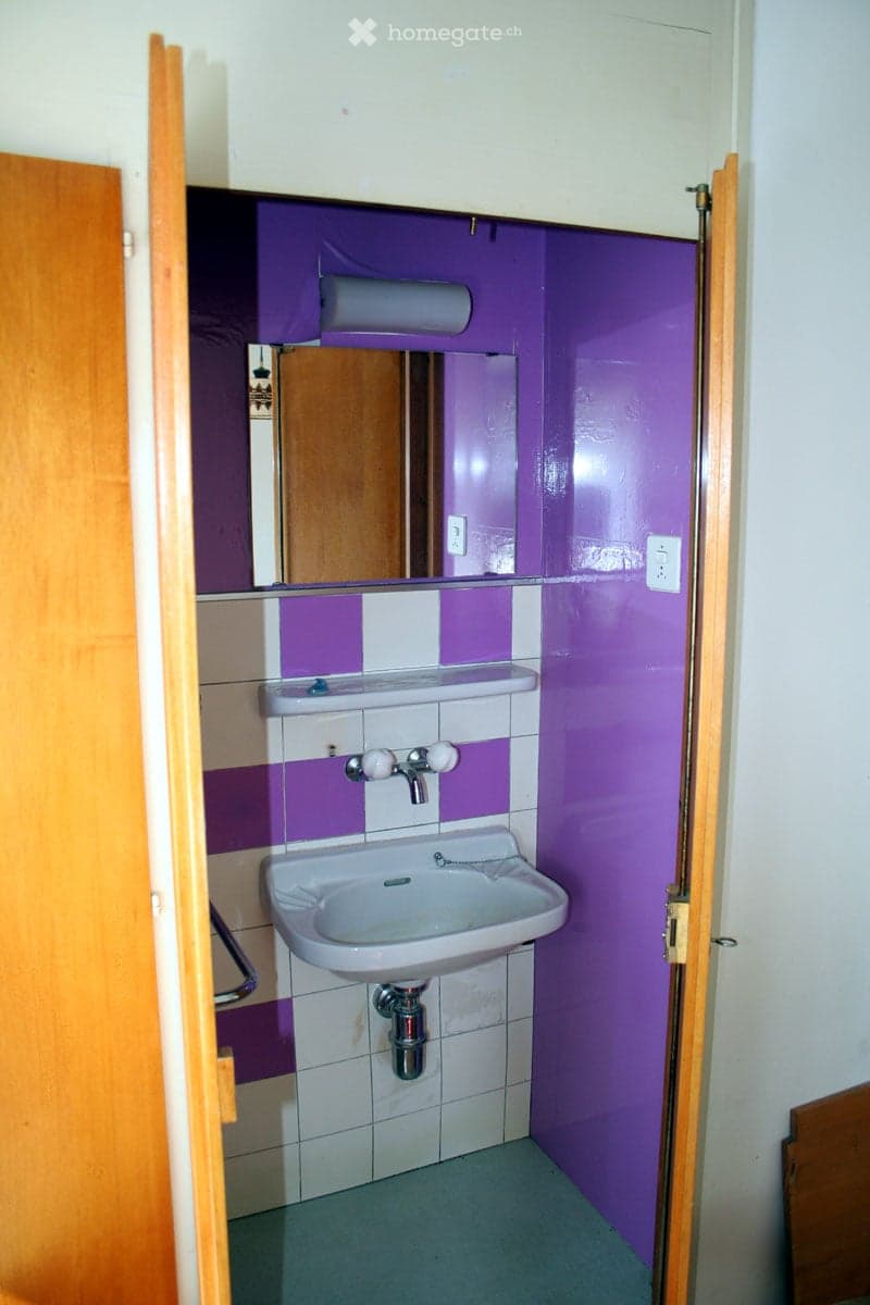 Small bathroom with purple walls, white tiled lower half, a single sink with a mirror above it, and a toilet paper holder on the wall.