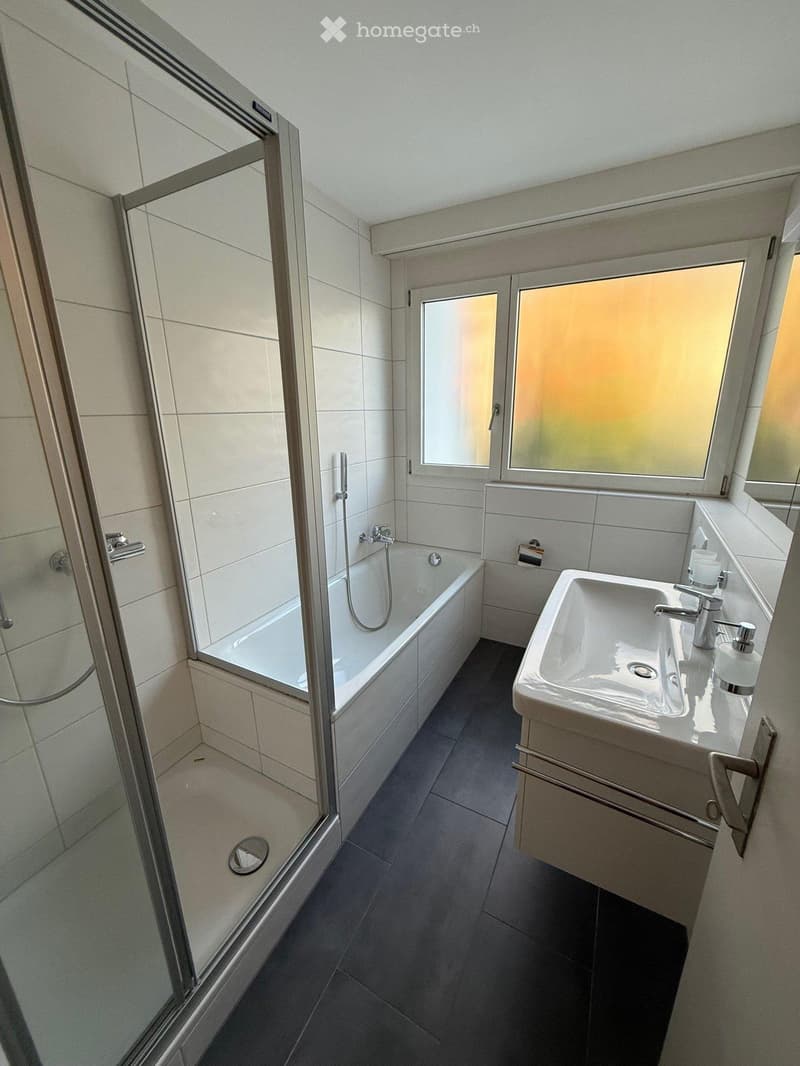 Modern bathroom with a bathtub, glass-enclosed shower, sink, and two windows. The space features white tiles and dark flooring.