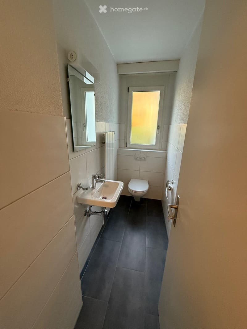 Compact bathroom with a white sink, chrome faucet, toilet, and a small mirror above the sink. The room has light-colored tiles and a dark floor.