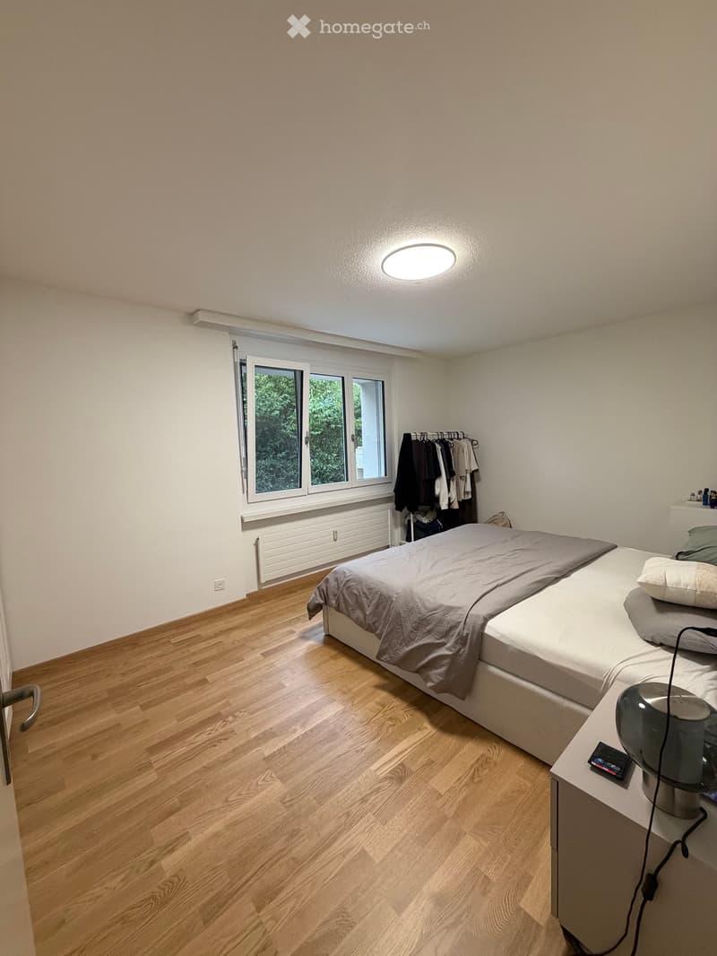 Cozy bedroom with wooden flooring, a double bed with grey bedding, a nightstand with a lamp, and a clothing rack with hanging garments.