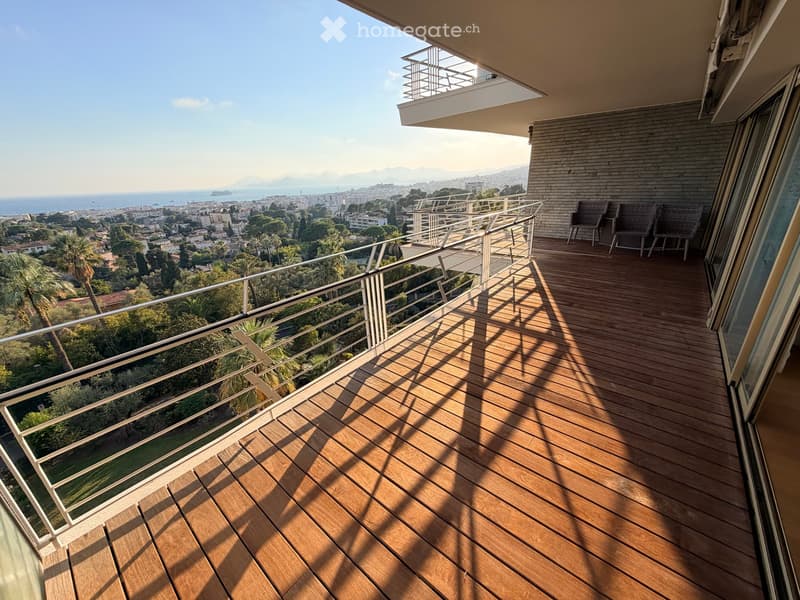 Wooden decked balcony with metal railings, overlooking a scenic coastal cityscape with the sea and distant mountains.