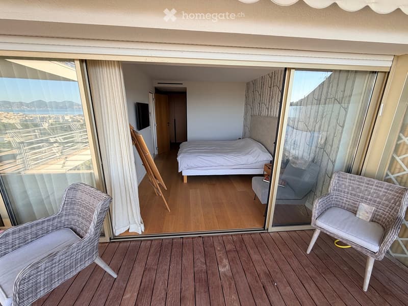 A furnished room with a bed, a TV, a small table, and two wicker chairs on a wooden deck, with a scenic view of the city and sea.