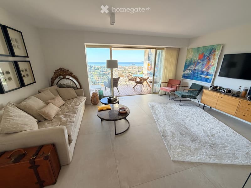 Modern living room with a beige sofa, coffee table, large windows offering a sea view, and a balcony with outdoor seating.