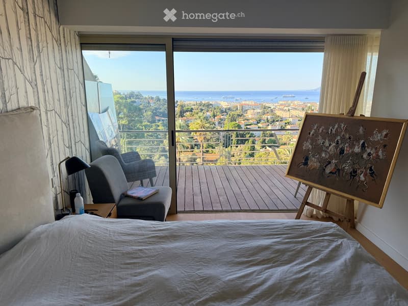 Bright bedroom with a large bed, a sofa, a chair, a small table with a lamp, and a painting on an easel. Large windows offer a scenic view of the sea and cityscape.