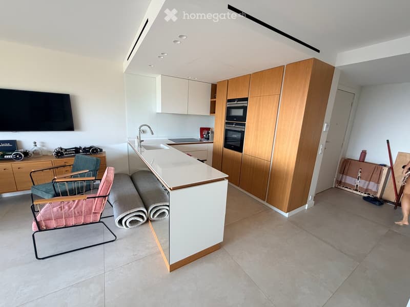 Modern kitchen with white cabinets, wooden accents, and a central island. Includes a sink, faucet, oven, microwave, and two chairs near a small table. There is also a screen mounted on the wall and some decorative items on a sideboard.
