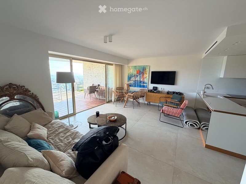 Modern living room with a sofa, coffee table, chairs, dining table, television, and a kitchen area with a counter and sink. Large windows offer a view of the ocean.