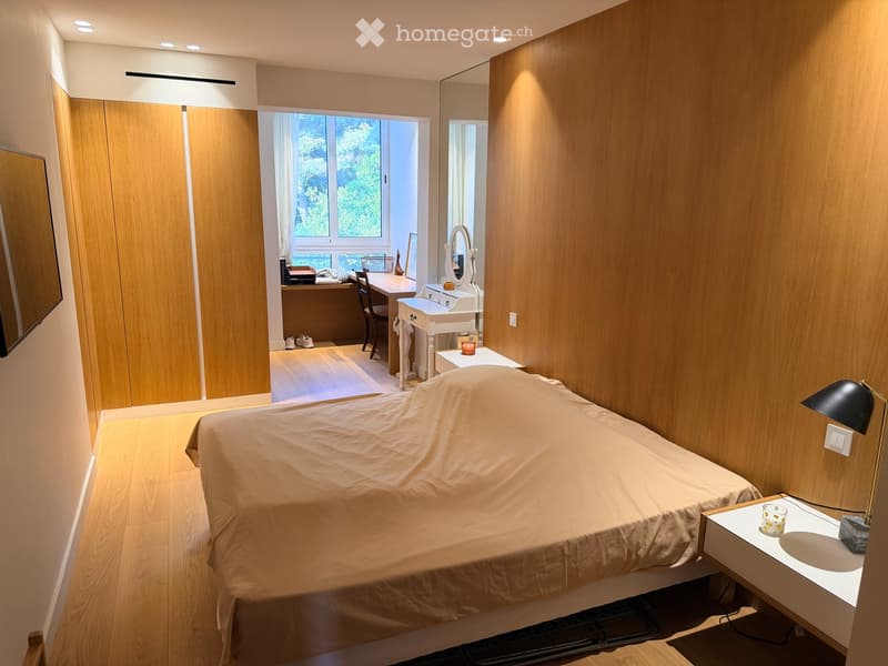 Modern bedroom with a large bed, wooden floor, built-in wardrobe, desk with chair, and a small sink area with a mirror and lamp.