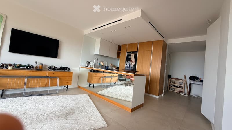 A modern, open-concept living and dining area with a kitchen. The space features a large television mounted on the wall, a sleek kitchen with white and wooden cabinets, a central island with bar stools, and a cozy living area with a plush white rug and a