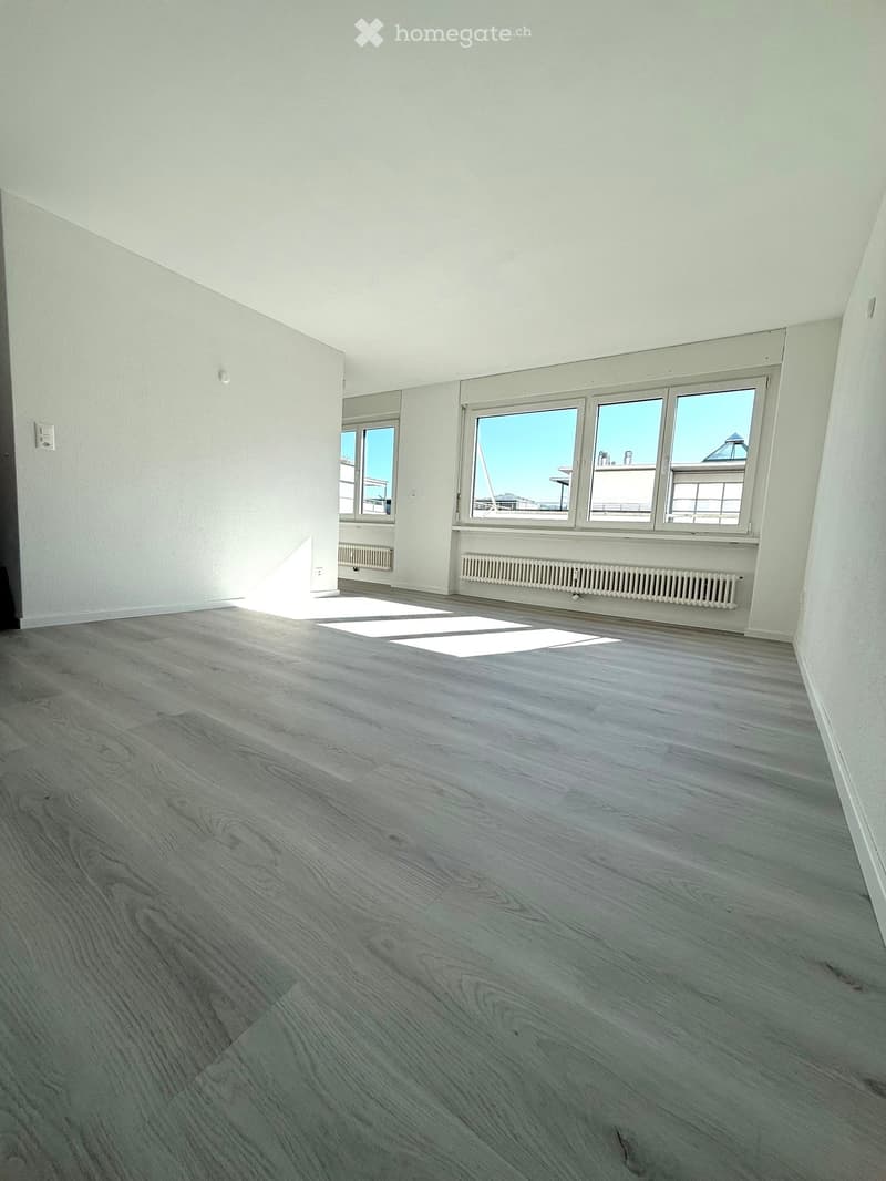 Empty room with light grey wooden flooring, white walls, and large windows offering a view of the cityscape.