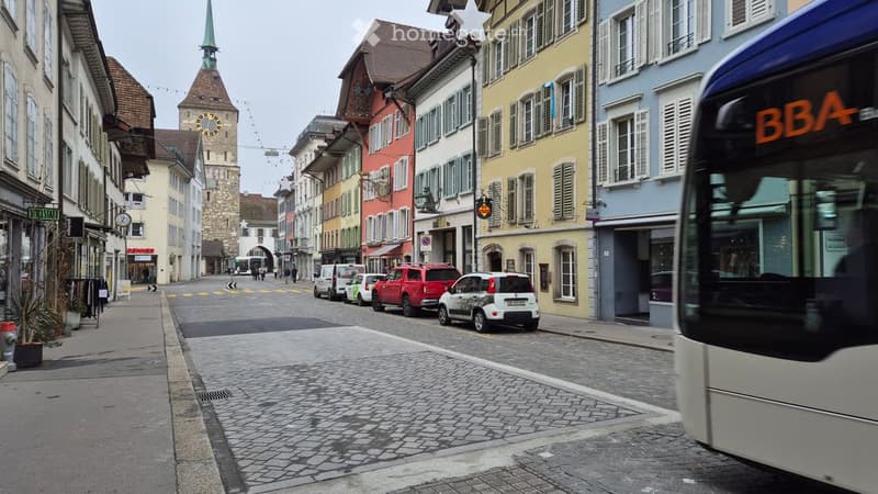 A European town square with historic buildings, a clock tower, and a bus passing by.