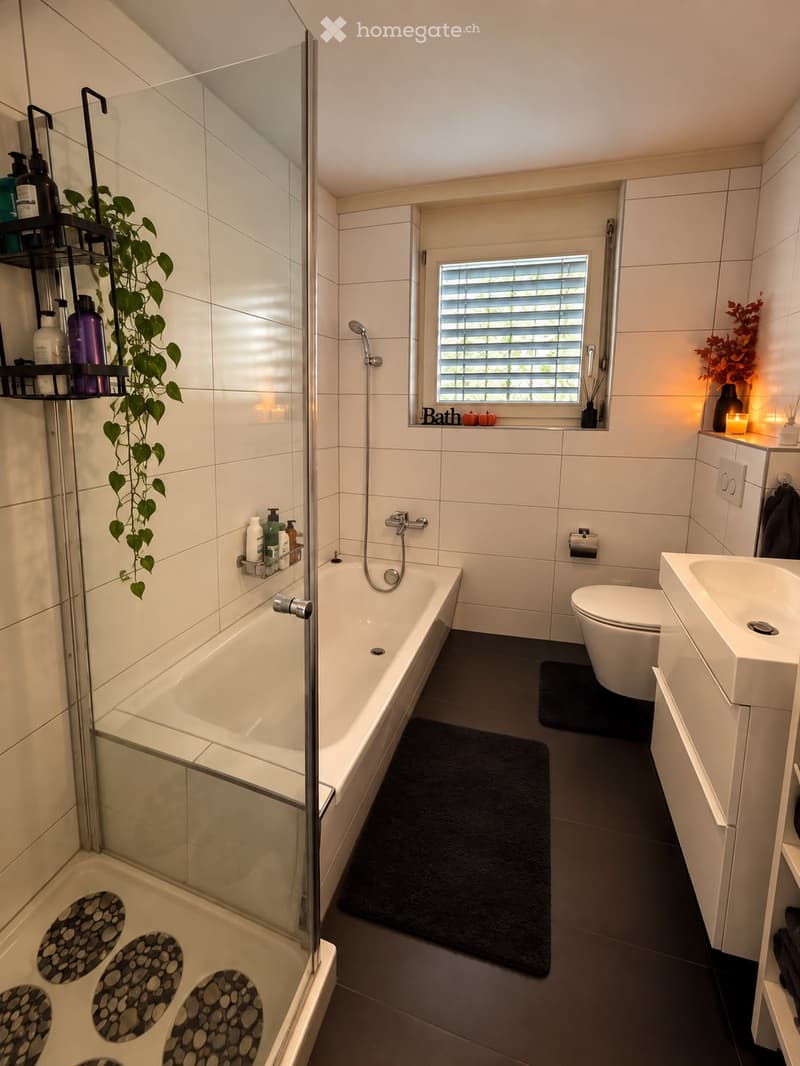 Modern bathroom with a bathtub, toilet, sink, and shower area. Features a black bathroom rug, a plant on a shelf, and a candle on the windowsill.