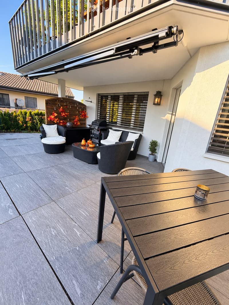 A modern outdoor patio with a wooden dining table, chairs, a wicker sofa set, a coffee table, a grill, and a small potted plant. The area features a tiled floor and is partially covered by a retractable awning.