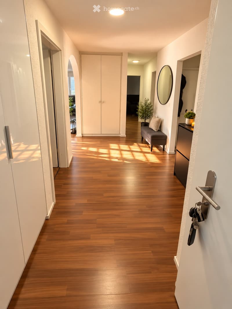 Bright hallway with wooden flooring, white walls, and recessed lighting. Features a bench with a cushion, a small table with a plant, and a round mirror on the wall.