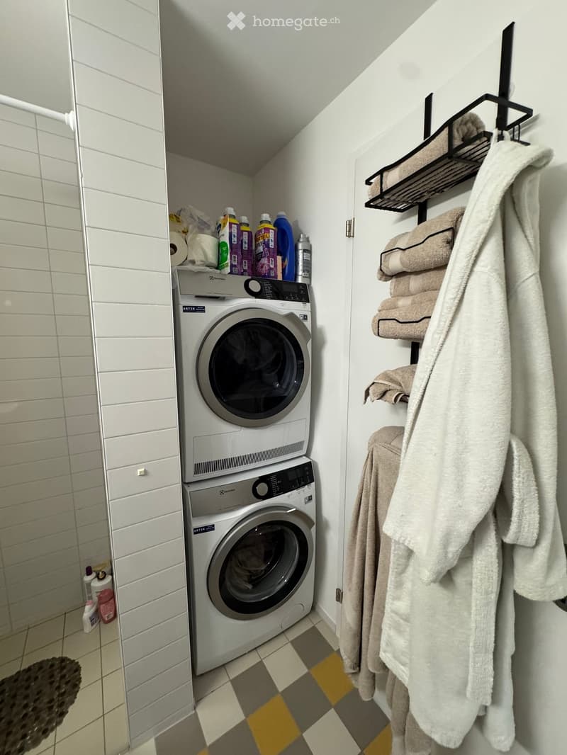 A compact bathroom with a stacked washing machine and dryer unit, a towel rack with neatly folded towels, and cleaning supplies on top of the washing machine.