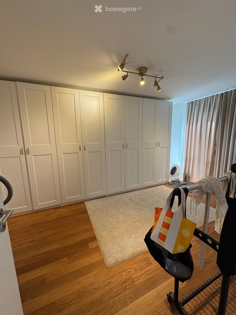 A well-lit room with wooden flooring, large white wardrobe, a small rug, and a clothes rack with a handbag and other items.