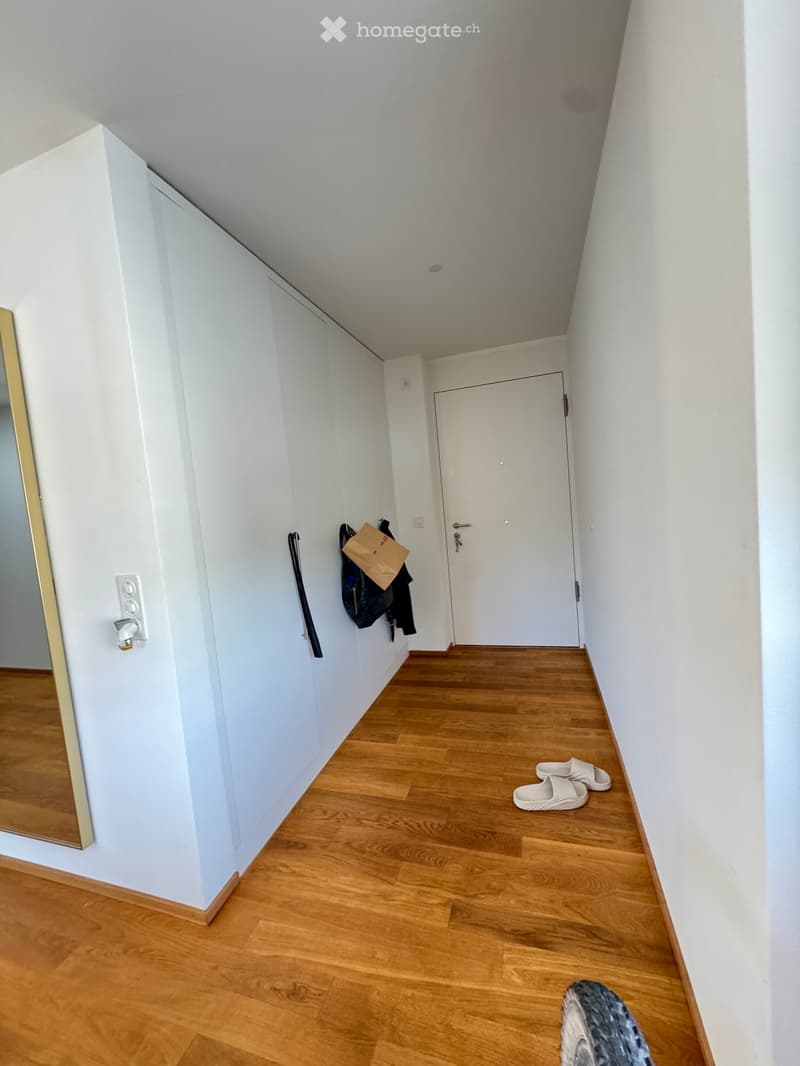 A narrow hallway with light wooden flooring, white walls, and a white door at the end. A coat rack with a jacket and a pair of slippers are visible.