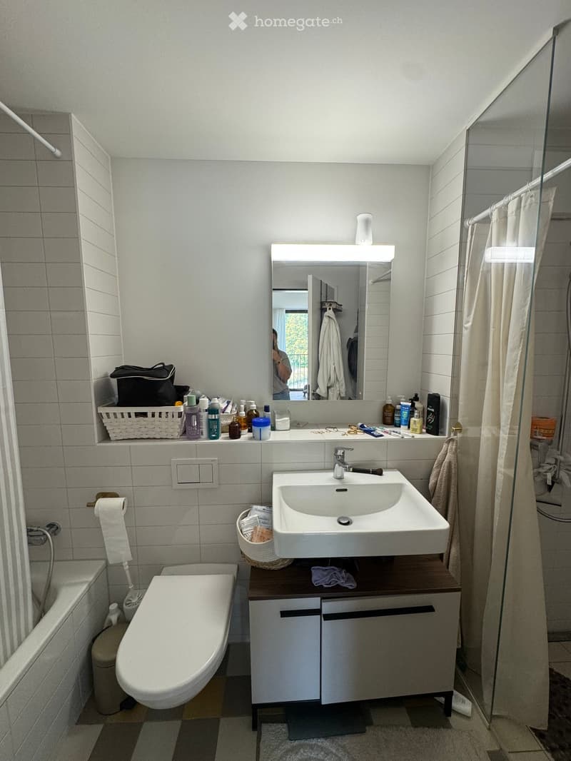 Modern bathroom with white tiles, a sink with a modern faucet, a mirror with built-in lighting, a toilet, a shower area with a curtain, and various toiletries on the counter.