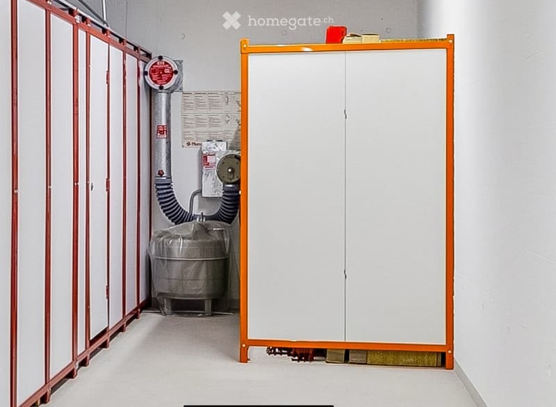 Empty utility room with white walls, grey floor, and a covered cylindrical object with a hose and control panel on the left side.