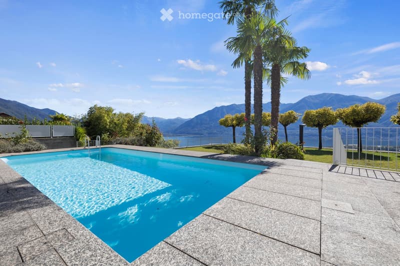 An outdoor area featuring a swimming pool with an entrance on the left, surrounded by a grey tiled floor. The pool has a metal handrail, and there are plants and trees nearby. There is a stunning view of mountains and a lake.
