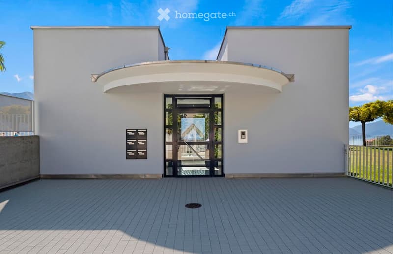 Modern white building, raised ground floor, entrance with a glass door, balcony, open courtyard, tiled floor, railings, outdoor greenery