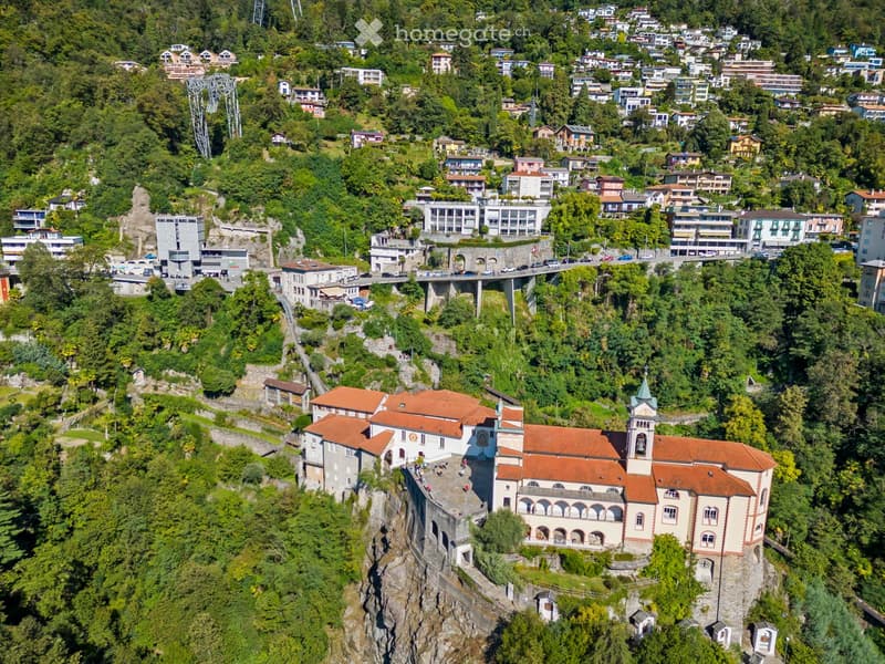 large historic building, white facade, red roofs, multiple windows, tower with belfry, on steep cliffside, surrounded by trees and shrubs, adjacent to a road and bridge