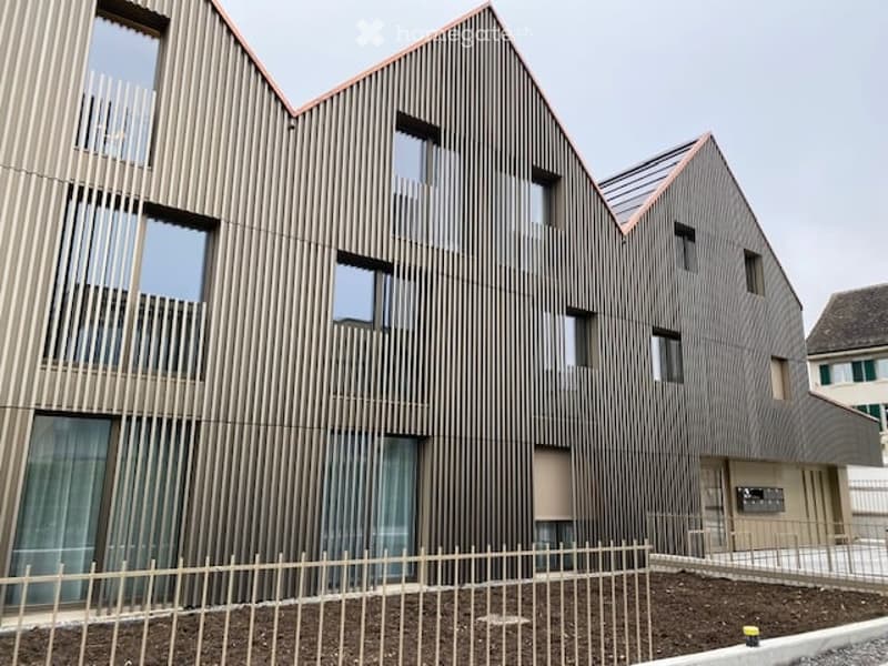 Modern building with wooden facade, multiple windows, metal fence, and a small garden area