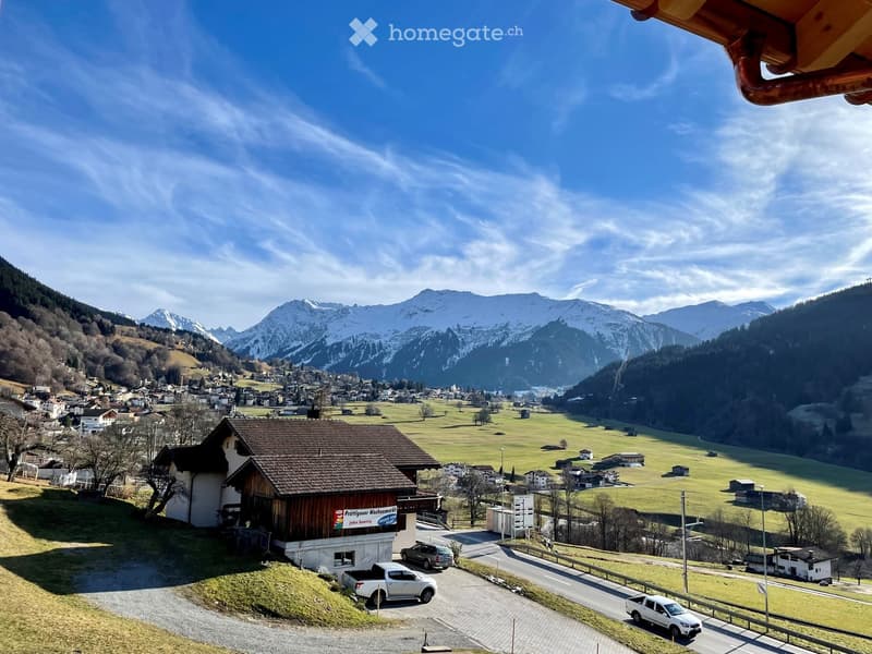 A scenic view of a small village nestled in a valley with snow-capped mountains in the background. The foreground shows a wooden building with a sign, a paved road, and several parked cars.