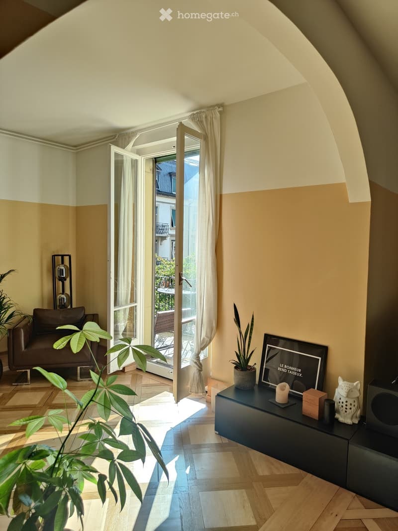 Bright living room with wooden flooring, large windows opening to a balcony, a brown leather sofa, a dark blue side table with a plant, a framed picture, and a candle.