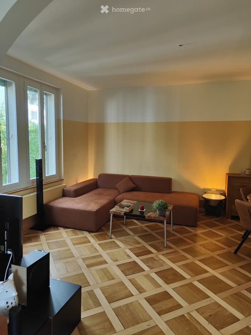Living room with wooden flooring, large window, brown sectional sofa, coffee table with a small plant, and a chair.